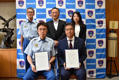 「秦野市消防本部」のロゴがある背景パネルの前で、制服姿の男性2人とスーツ姿の男性2人、女性1人の計5人が並び、前列に座る2人が白い書類を手に持っている締結式での写真