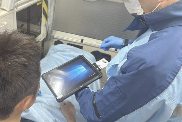 救急車内で、マスクと手袋を着用した救急隊員が、患者の横でタブレット端末に接続されたカードリーダーにマイナンバーカードをかざして読み取っている活動中の様子の写真