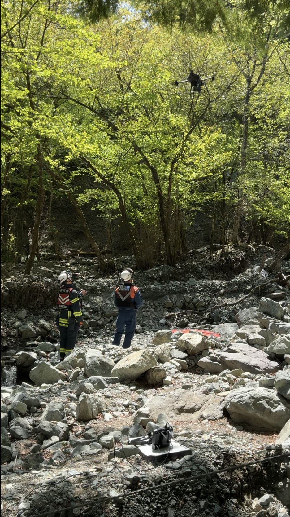 若葉が生い茂る山あいの岩だらけの川原で、救助服を着てヘルメットをかぶった二人の隊員が立っており、上空では小型のドローンが飛行しながら捜索または状況確認を行っている様子を捉えた写真