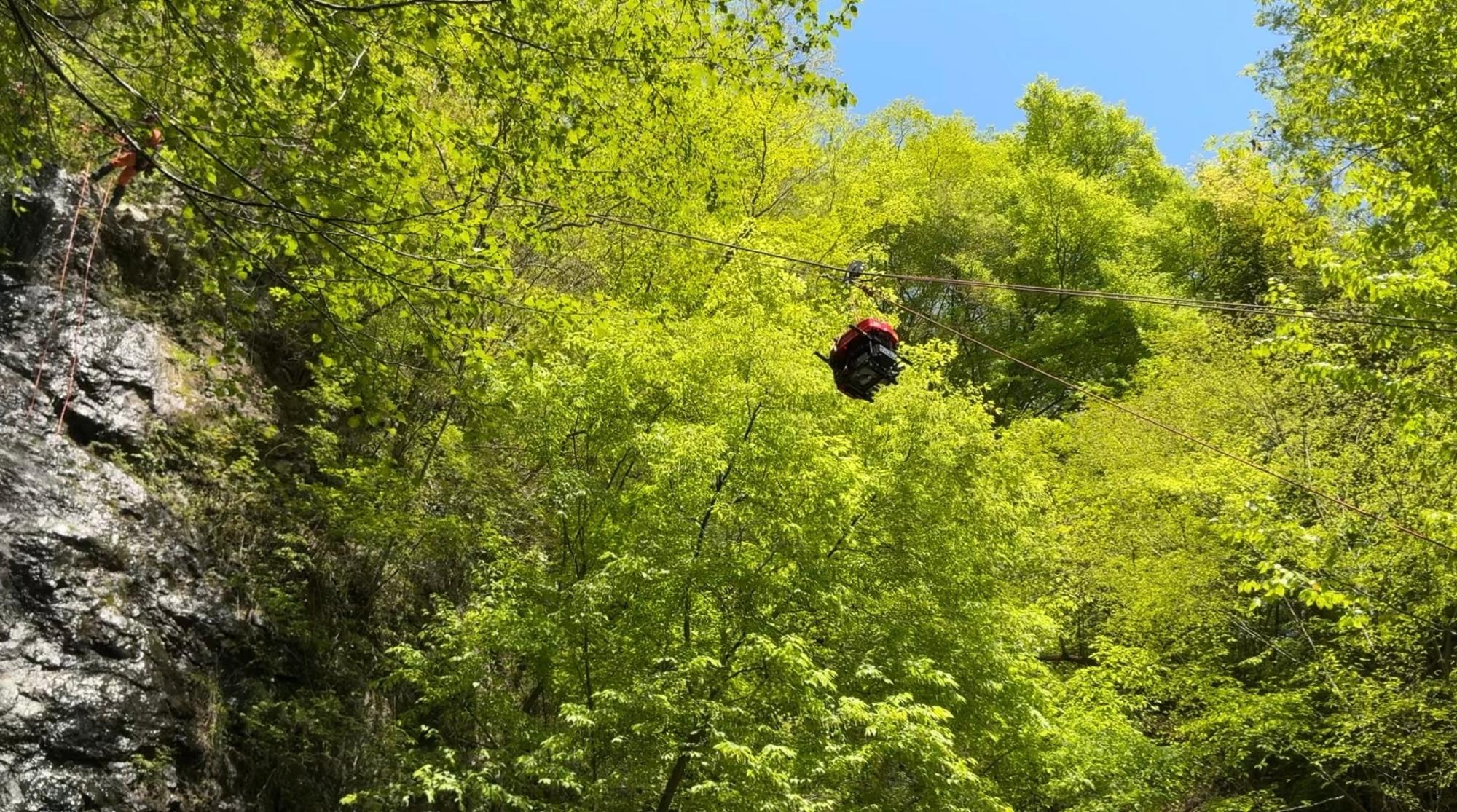 鮮やかな新緑の木々に囲まれた山間で、中央のロープウェイのようなワイヤーには黒い荷物が吊るされ滝の上部へ引き上げられている写真