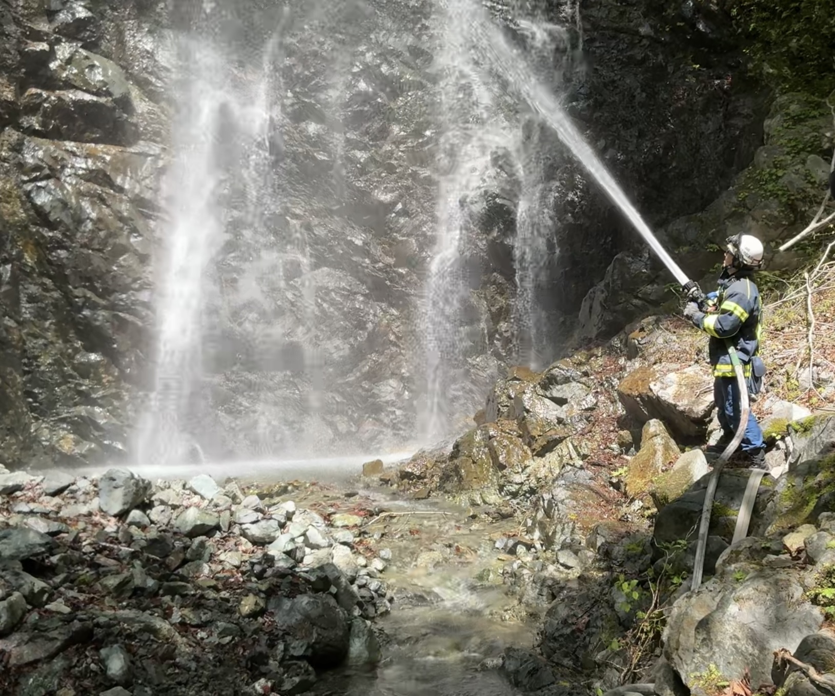 ゴツゴツとした岩場に囲まれた滝つぼ付近で、ヘルメットと防火服を着用した消防隊員が、放水ノズルから勢いよく水を放水し、水煙が立ち込める中で訓練を行っている様子の写真