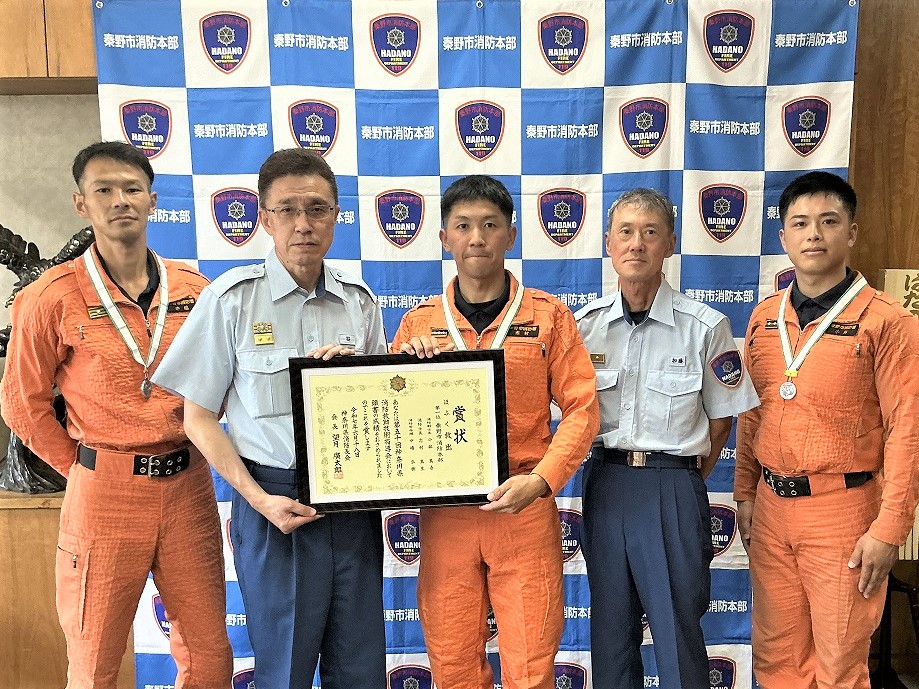 秦野市消防本部のバックボードの前でレスキュー服の男性3人と制服姿の男性2人が横並びになり、中央の2名が賞状を一緒に持っている記念写真