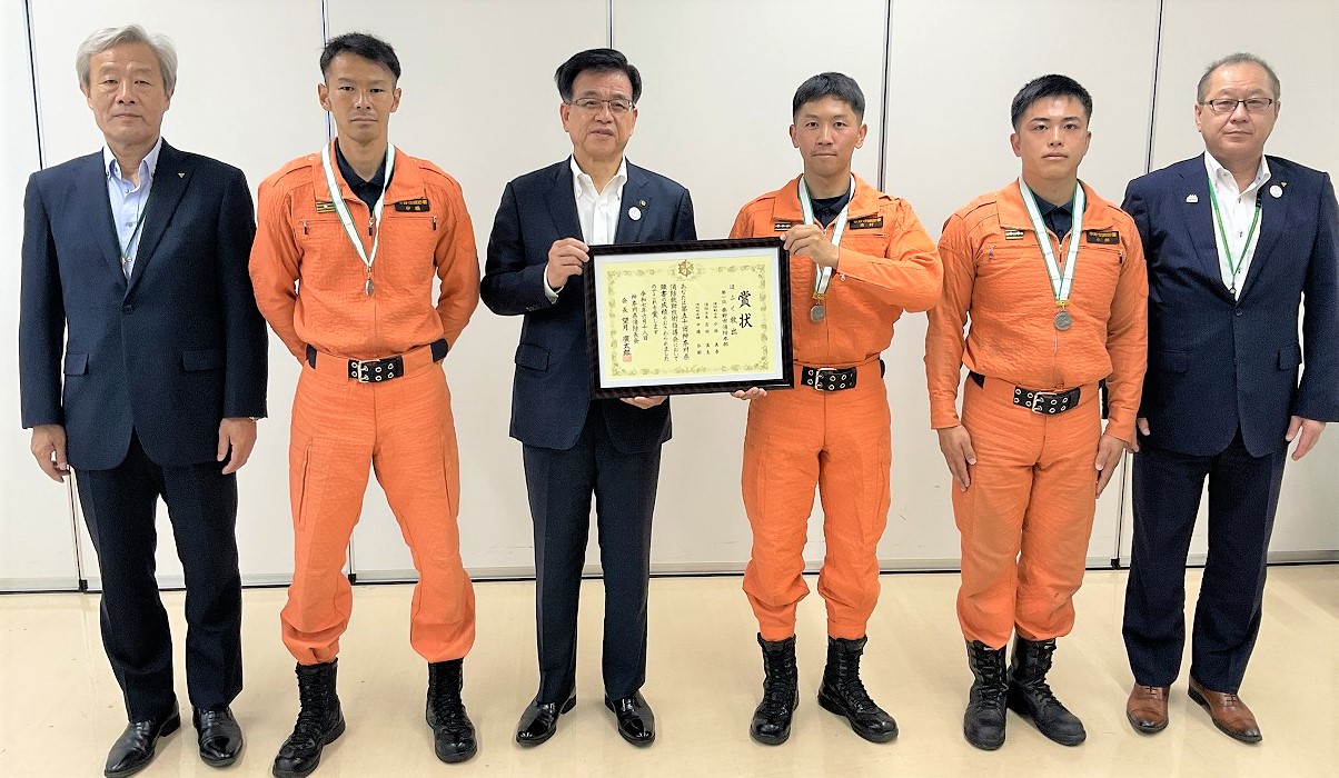 オレンジ色のレスキュー服と黒いブーツを着用した男性3名とスーツ姿の男性3名が横並びに並び、中央ののレスキュー隊の男性と市長が一緒に賞状を持ち記念撮影した写真