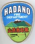 青空と緑の山々、雪を被った山が描かれ、上部に「HADANO FIRE DEPARTMENT」、中央下部に「山岳救助隊」の文字、ピッケルがデザインされた刺繍の山岳救助隊ワッペンの写真