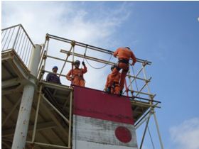 救助訓練用の櫓の上からオレンジ色の救助服を着た隊員の一人がロープで降下を始める様子を、下から見上げるように捉えた懸垂降下訓練の写真