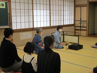 和室で着物を着た女性が正座し、茶道の作法を参加者に教えている様子を写した写真