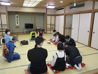 和室の畳の上で、着物を着た先生と複数の参加者が正座をして、茶道の体験会に参加している様子の写真