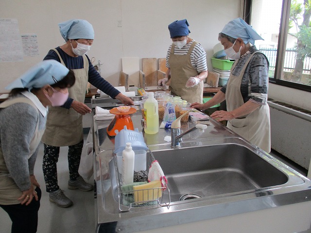 エプロンと三角巾をつけた女性4人が、調理室の調理台で材料や調味料を使い、調理作業を進めている様子の写真