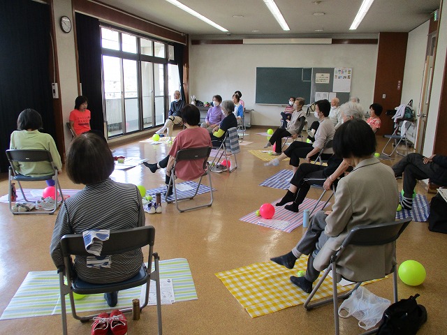 室内の広い空間で複数の参加者が椅子に座り、カラフルな敷物と柔らかいボールが足元に置かれ、健康維持のための体操や運動に参加している様子の写真