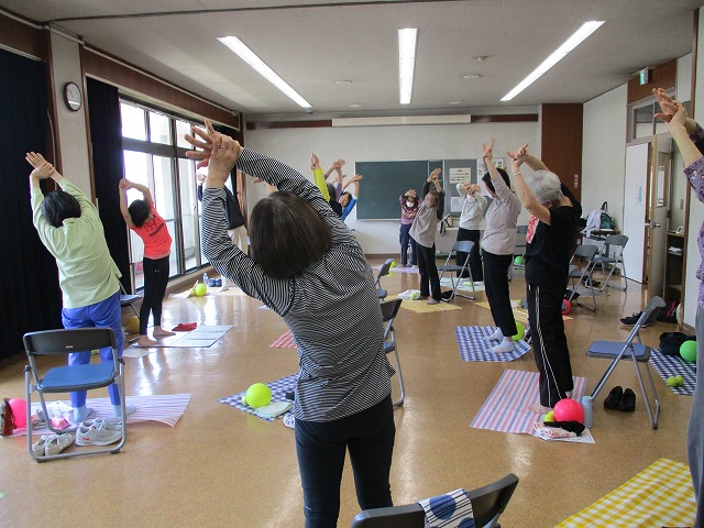 広い室内で、複数の参加者が両手を上げて体を横に曲げるストレッチをしている様子の写真