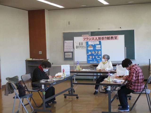 「フランス人形折り紙教室」と書かれ、折り方の説明などが貼られたホワイトボードの前でマスクを着けた女性3人が長机を囲んで折り紙をしている写真