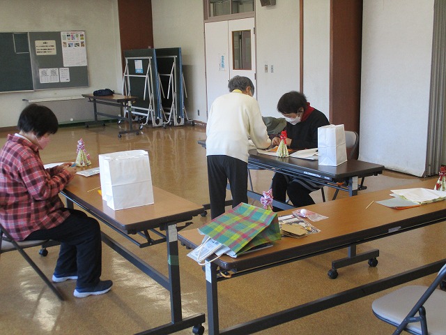 長机に座った2人の女性が折り紙をしており、もう1人がその様子を見て教えている写真