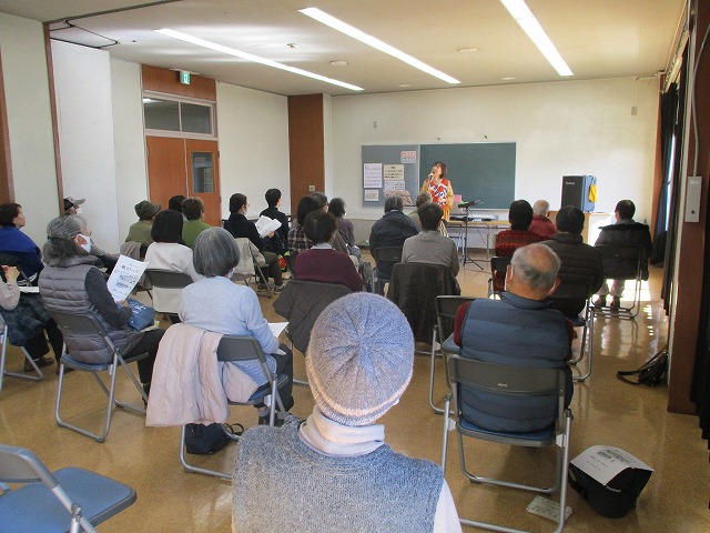 室内で複数の参加者が椅子に座り、前方でマイクを持った女性の教師の話を聞いているのを後方から写した写真