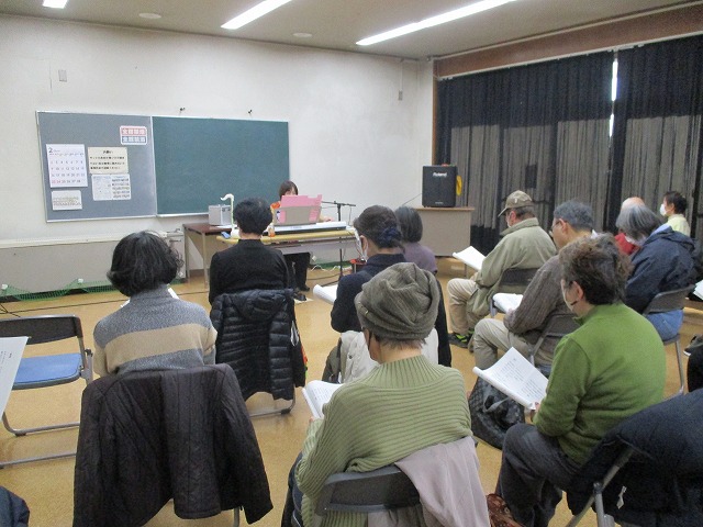 室内で、ピアノを弾く講師の前方に、多数の参加者が椅子に座り、歌詞を見ながら聴講している様子の写真