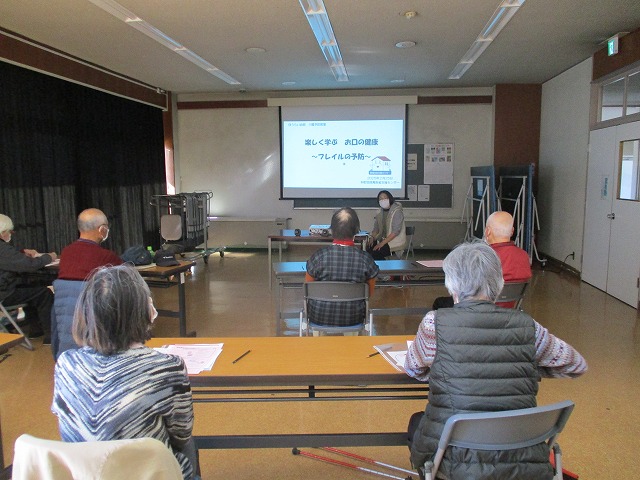 複数の参加者がスクリーンに映し出された「楽しく学ぶ お口の健康」というスライドを見ながら、講師の話を聞いているのを後方から写した写真