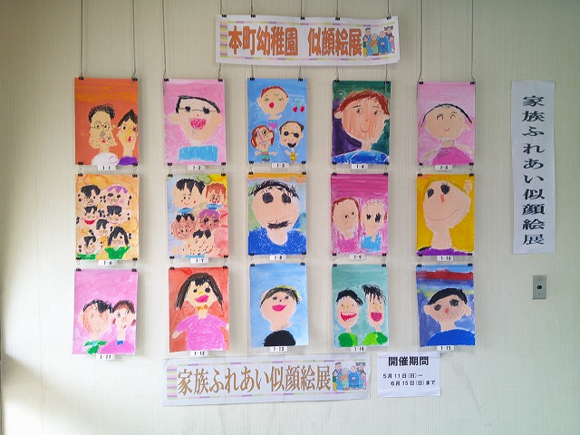 幼稚園児が描いた家族の似顔絵が縦4列&times;横4列で壁に飾られており、上には「本町幼稚園 似顔絵展」と書かれ、下には「家族ふれあい似顔絵展」と表示がある写真