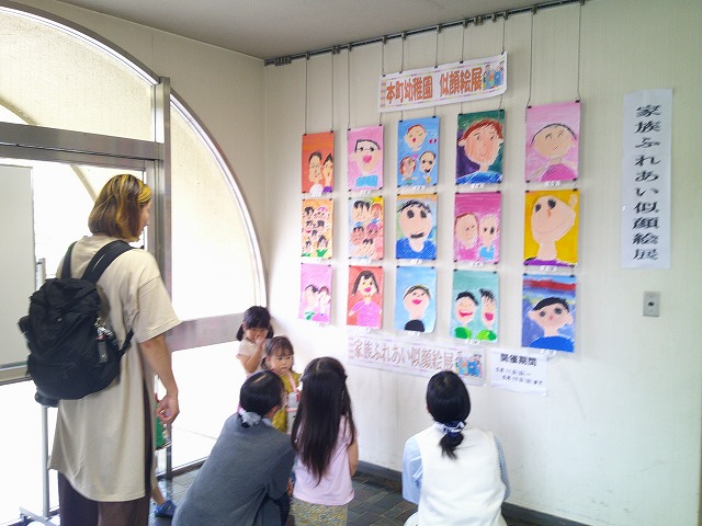 子どもと保護者数人が、壁に飾られた幼稚園児の似顔絵を見上げたり、しゃがんで鑑賞したりしている様子の写真
