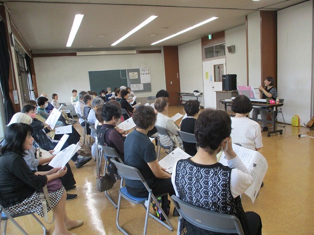 多くの方が集会所で椅子に座り、歌詞を見ながら、前方の女性講師の指導を受けている写真