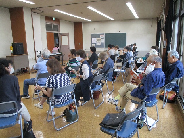 前方にはキーボードとマイクがあり、高齢者を中心とした男女が、椅子に座って参加者が資料を見たり話をしている室内の様子の写真