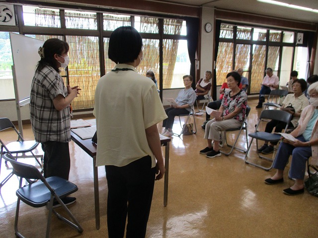 講師の女性が前に立ってマイクで説明をし、参加者の高齢者たちが資料を持ち椅子に座って話を聞いている写真