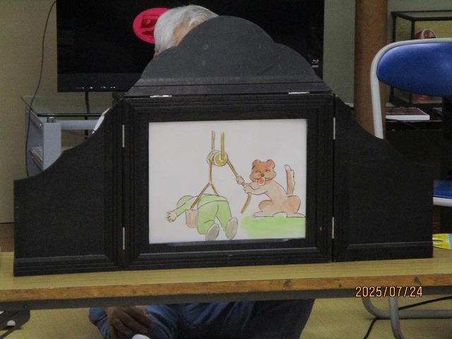 井戸に落ちた人を動物が助けようとしている絵の紙芝居が、木製の舞台を使って演じられている様子の写真