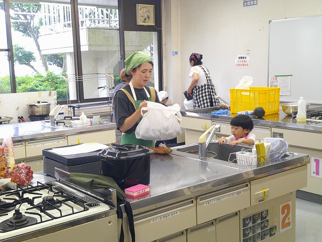 調理実習室で、エプロン姿の女性が準備をし、傍らの流し台では幼い子供が顔を覗かせている微笑ましい様子の写真