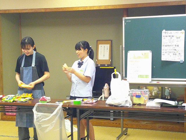 公民館の和室で、制服姿とエプロン姿の2人の女子学生が、テーブルにお菓子などを広げてイベントの準備をにこやかに進めている様子の写真