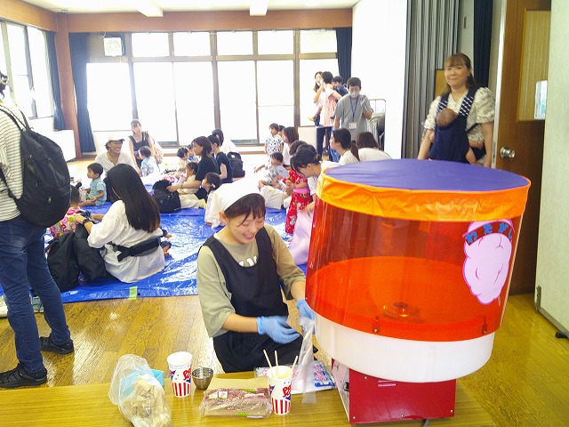 室内イベントで、ブルーシートに座る大勢の参加者を前に、エプロン姿の女性がわたあめ機でわたあめを作っている様子の写真