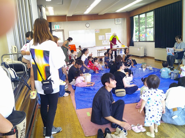 大勢の親子がブルーシートに座る室内イベントで、絵本の読み聞かせや前の舞台での出し物を楽しんでいる様子の写真