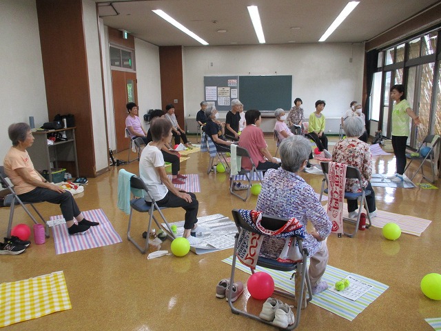 カラフルな敷物と柔らかいボールが使われ、椅子に座った高齢者たちが指導者の説明を受けながらゆっくりと体操をしている写真