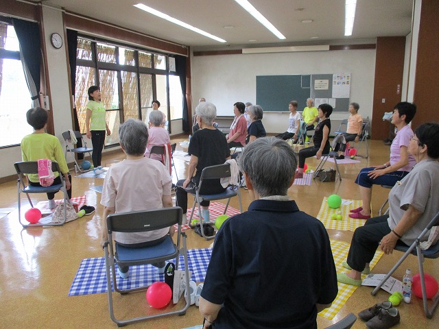 明るい室内で高齢者たちが椅子に座って姿勢を整え、前方の講師の指示に従って軽い運動をしており、タオルやボールなども活用している椅子体操の写真
