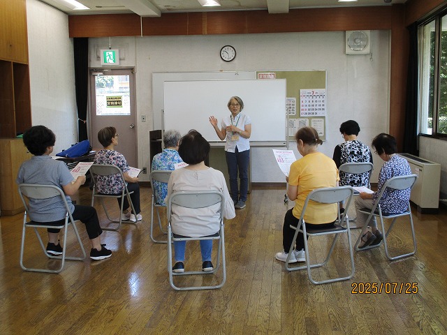 教室のような部屋で講師の女性が立って話し、数人の高齢の女性たちが椅子に座ってプリントを持ちながら講義を受けている写真