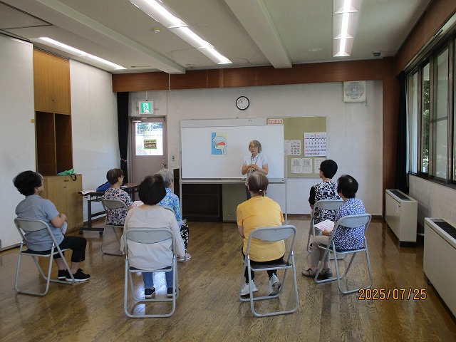 ホワイトボードに口や喉の図が貼られた前で講師の女性が説明しており、参加者の高齢女性たちが椅子に座って真剣に話を聞いている講習会の写真