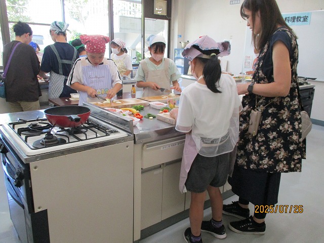 調理室で子どもたちがエプロンや三角巾をつけて包丁で野菜を切るなど料理をしており、大人が横で見守っている写真