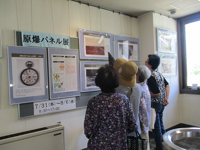 「原爆パネル展」と書かれた展示の前で、高齢の女性たちが戦争に関する写真や資料パネルを真剣に読みながら鑑賞している写真