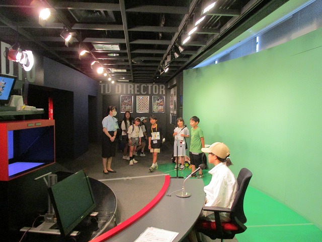 テレビスタジオ風の部屋でアナウンサー役の子が机に座ってマイクの前で話し、子どもたちの横で関係者の方が説明している写真