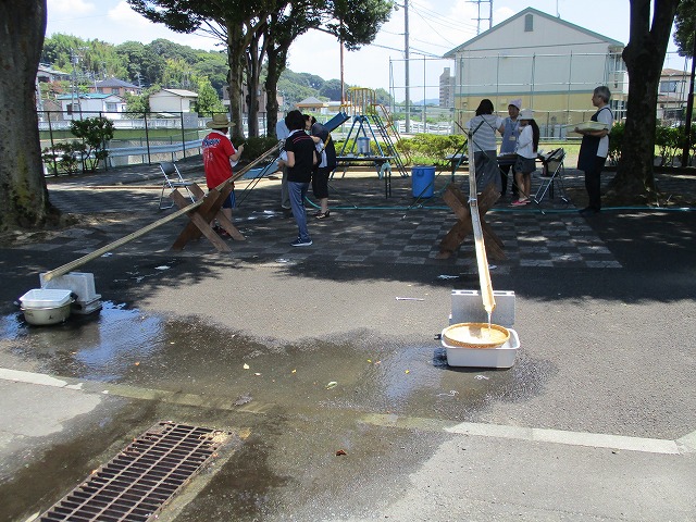 長い竹を半分に割って流しそうめんの装置を作り、子どもたちと大人が水を流しながらそうめんを取って楽しんでい楽しんでいる様子で、背景には公園の遊具と木陰が見える流しそうめん体験の写真