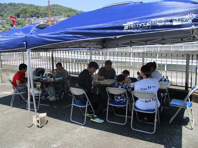 青いテントの下に複数のテーブルと椅子が並べられ、家族連れや子どもたちが集まって食事を楽しんでいる屋外交流の写真