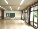 大きな窓から自然光が入り、蛍光灯が天井に並んだ広いフローリングの部屋で、壁に黒板がかけられている集会室の写真