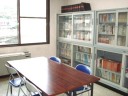 大きな窓から光が差し込む空間で、木製の長机と青い椅子が配置され、壁際にはガラス扉の書棚に多くの書籍や資料が収納されている写真