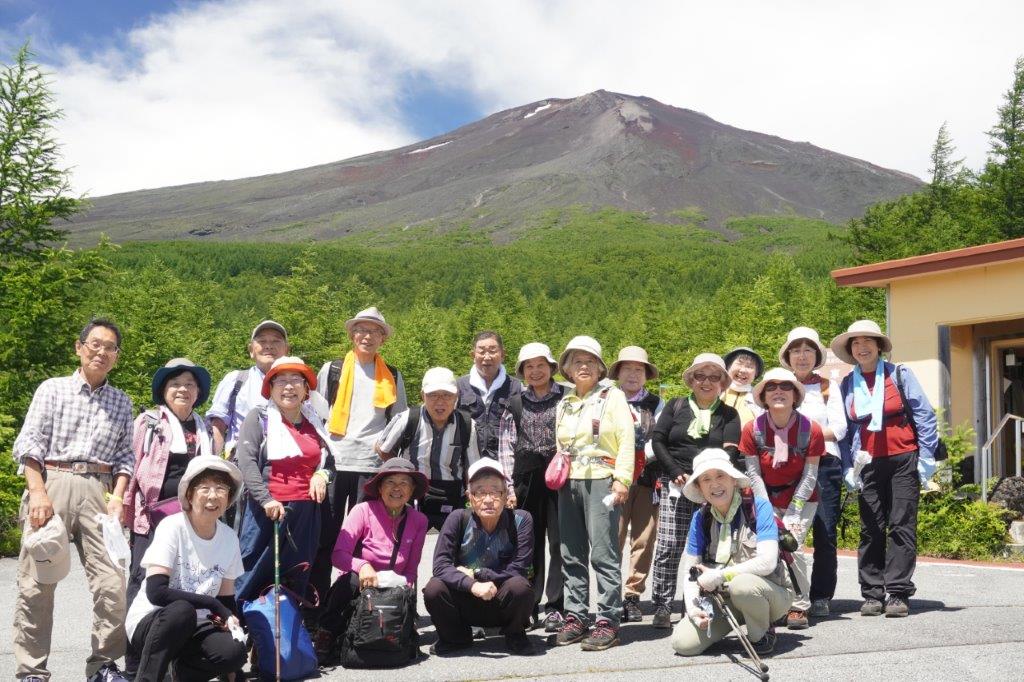 快晴の空の下、多数の高齢者がハイキングウェアや帽子を身に着けて、富士山のような雄大な茶色い山と緑豊かな針葉樹林を背景に、楽しそうな表情で集合している登山グループの記念写真