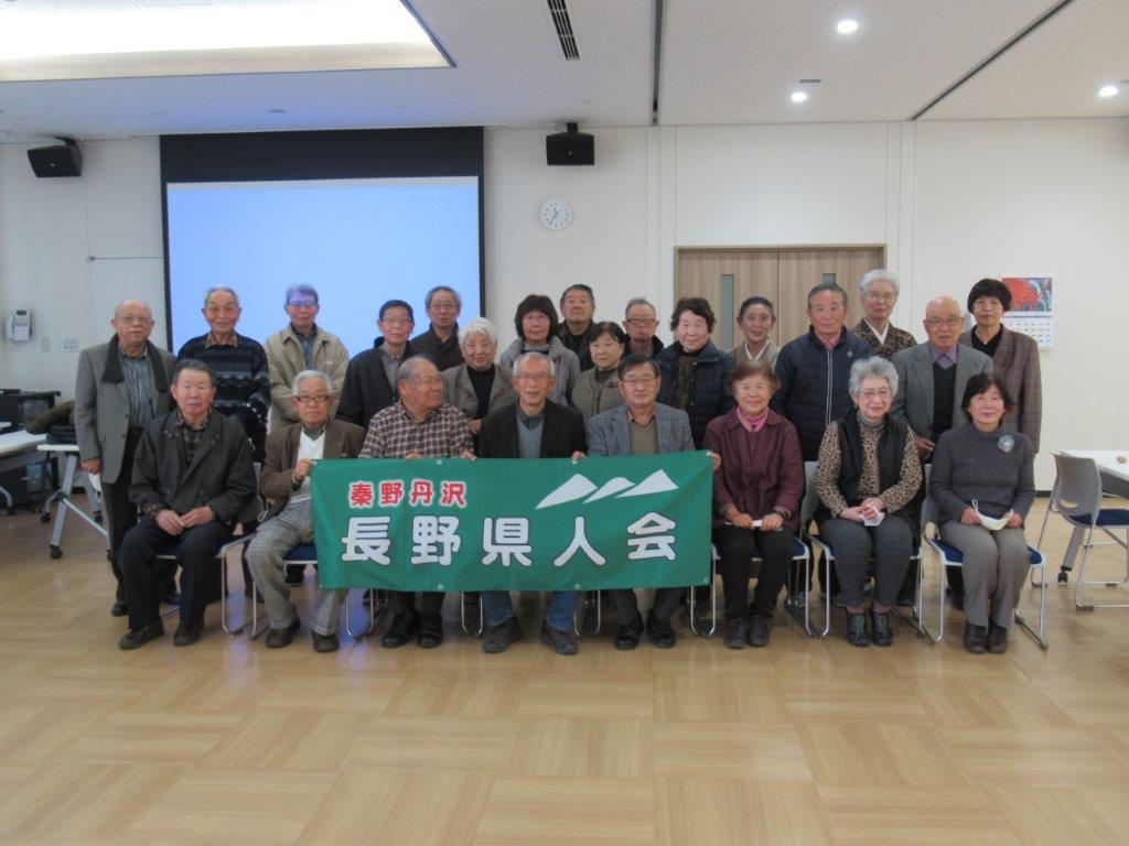 屋内ホールのような広い会場で、多数の高齢者が、緑色の横断幕に「秦野丹沢 長野県人会」と書かれたものを掲げながら、二列に並んでカメラに向かって立っている、会議や集会後の記念撮影のような集合写真
