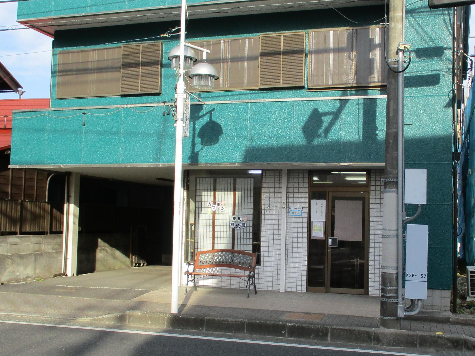 緑がかった青の壁が特徴的な、一階が駐車場と店舗の入り口になっている建物が、昼間の歩道から撮影されたあつまる本町交流館の外観写真