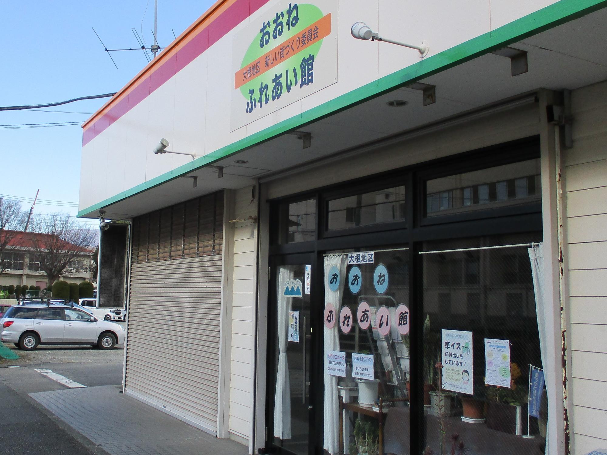 白い建物にオレンジと緑のラインが入った外観で、「おおね ふれあい館」と書かれた看板とガラス扉には「大橋地区 おおね ふれあい館」の文字が確認できる、建物の入口付近を斜めから捉えた写真