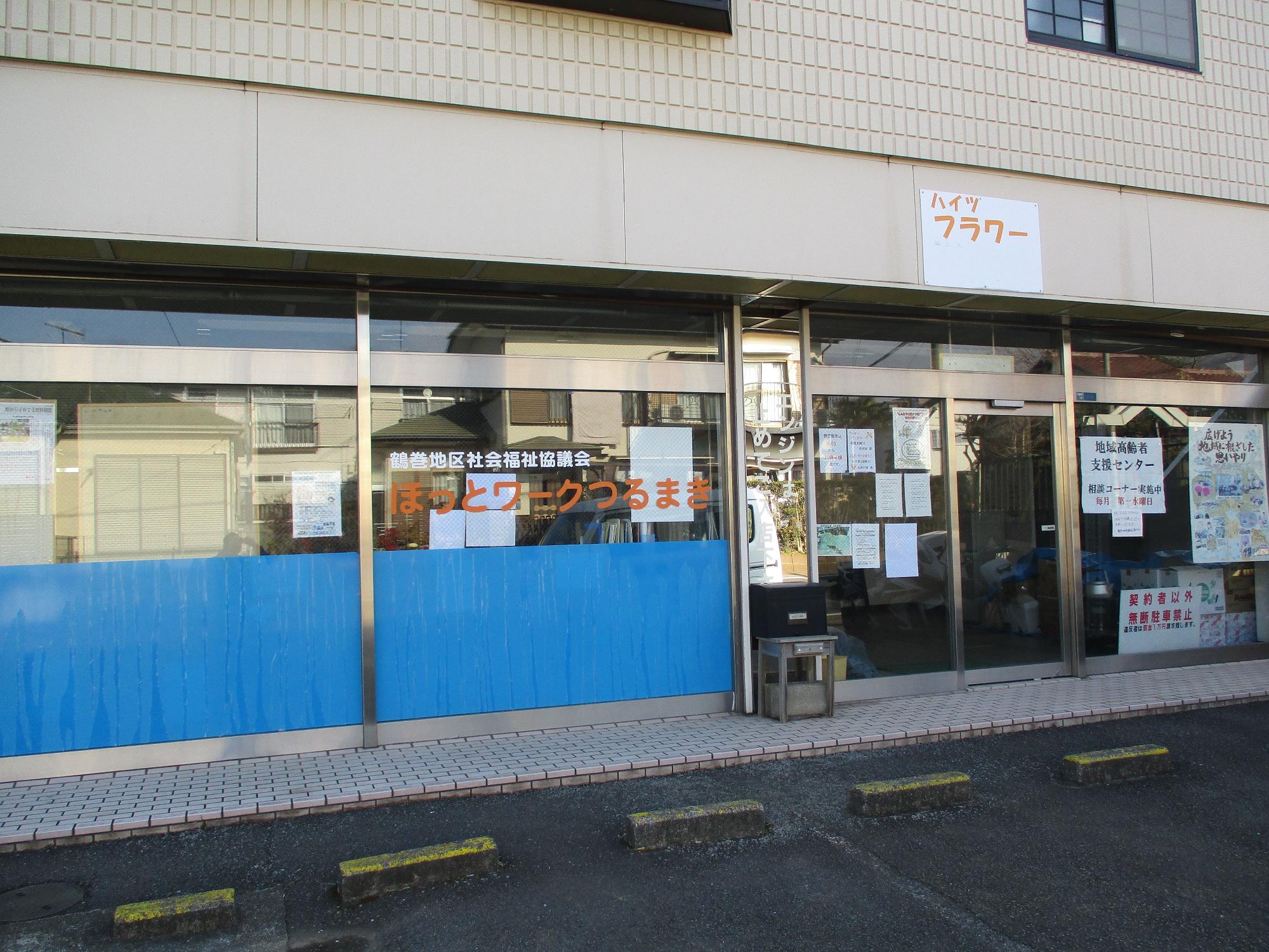 白っぽい壁の建物の一階に位置する施設で、大きなガラス窓には青いシートが貼られ、多くの掲示物が貼られている「福祉とワークつるまき」と書かれた建物の外観を、舗装された駐車場から捉えた写真