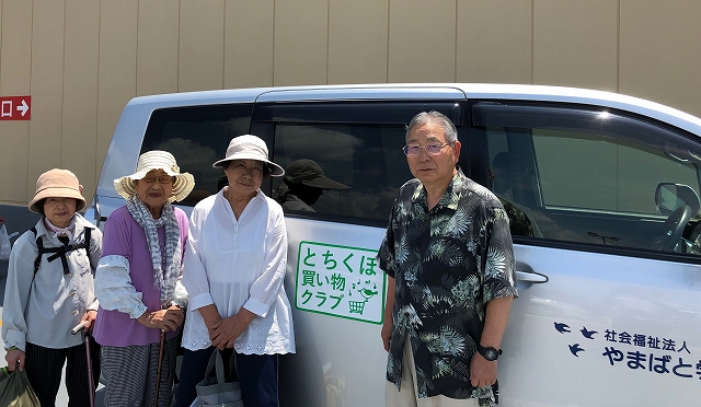 「とちぼ買い物クラブ 虹」と書かれたシルバーのワゴン車の横で、和装の上着を羽織った男性と麦わら帽子などをかぶった三人の高齢女性が並んで立っている写真