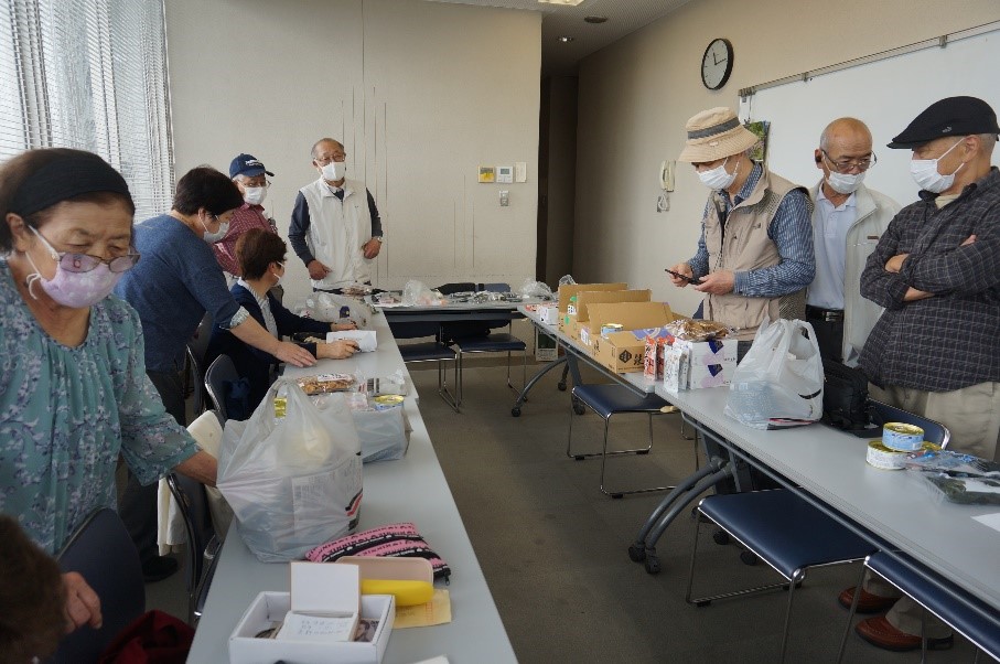 会議室らしき屋内のスペースで、マスクを着用した10人ほどの高齢者が、テーブルの上に並べられた多数の食品や日用品を仕分けたり、手に取って確認したりしている様子を写した写真
