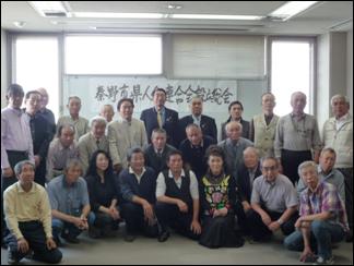 会議室のような明るい室内で、多くの高齢の男性と数名の女性が写っており、中央の白板には手書きで「秦野市退職者連合総会」のような文字が確認できる集合写真