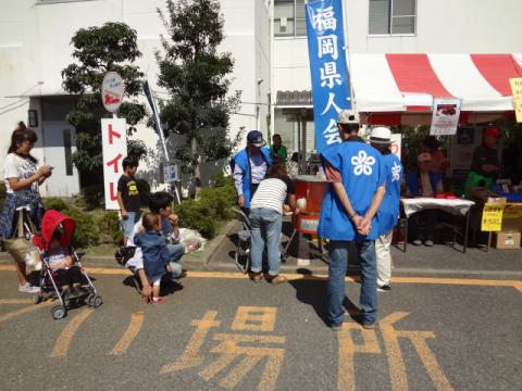 秋の青空の下、アスファルトの地面に「場所」と黄色い文字で書かれた場所の奥で、青い法被を着た男性たちが屋台の準備をし、その手前でベビーカーの赤ちゃんや子供連れの家族が賑わうお祭りかイベントの準備風景の写真