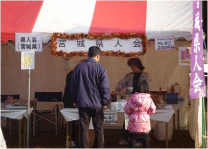 日差しを浴びた屋外イベント会場で、赤と白のストライプのテントの下にある「宮城県人会」のブースにおいて、客の男性と小さな女の子が、店員から温かい飲食物を受け取っている販売風景の写真